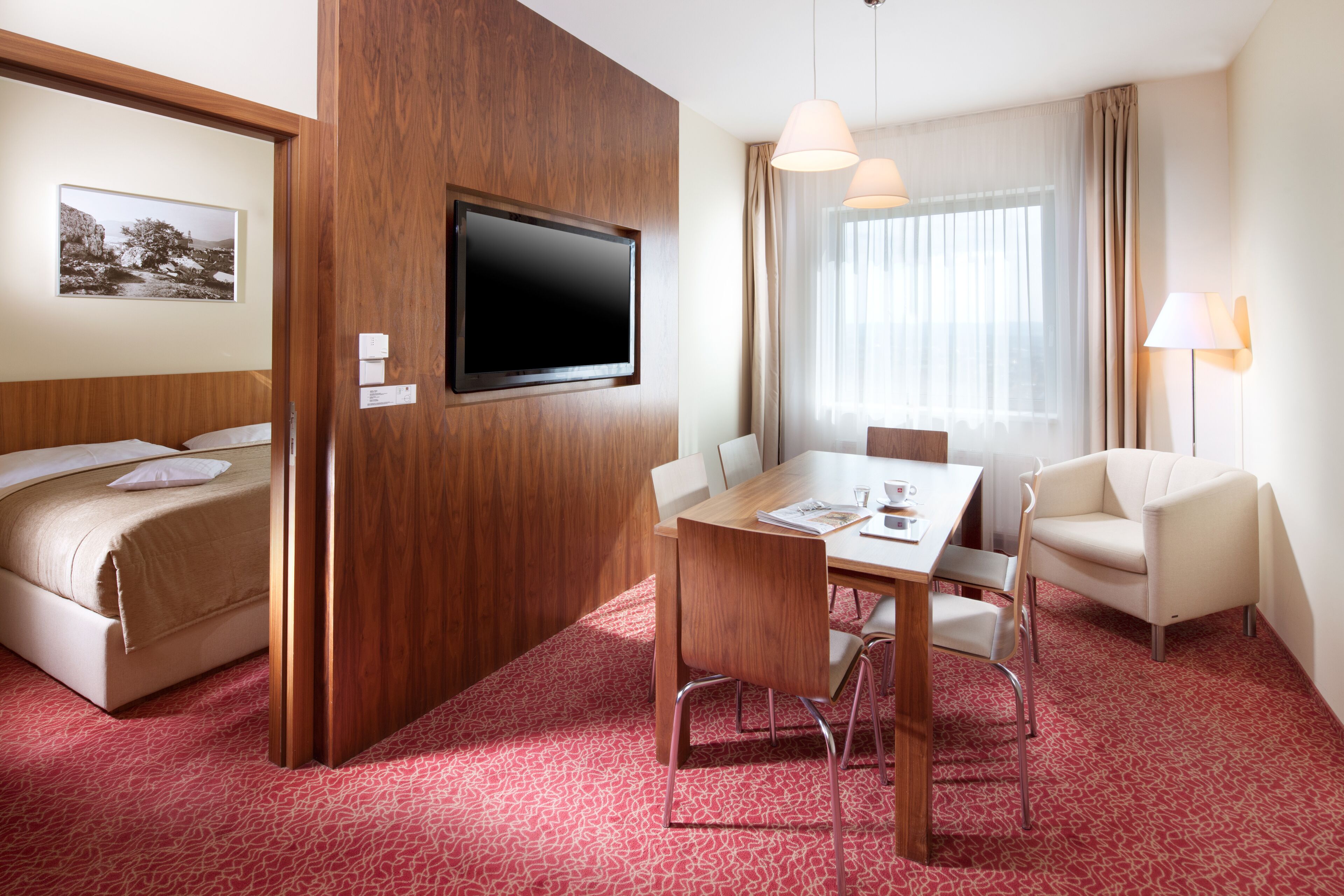 Ein Hotelzimmer mit Bett, Fernseher an einer Holzverkleidung, Esstisch mit Stühlen, Lampe und einem fenster mit Vorhängen.