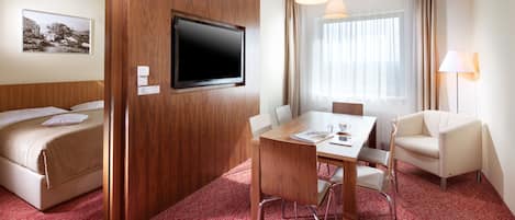 A hotel room with a bed, a TV mounted on a wooden panel, a dining table with chairs, a lamp, and a window with curtains.
