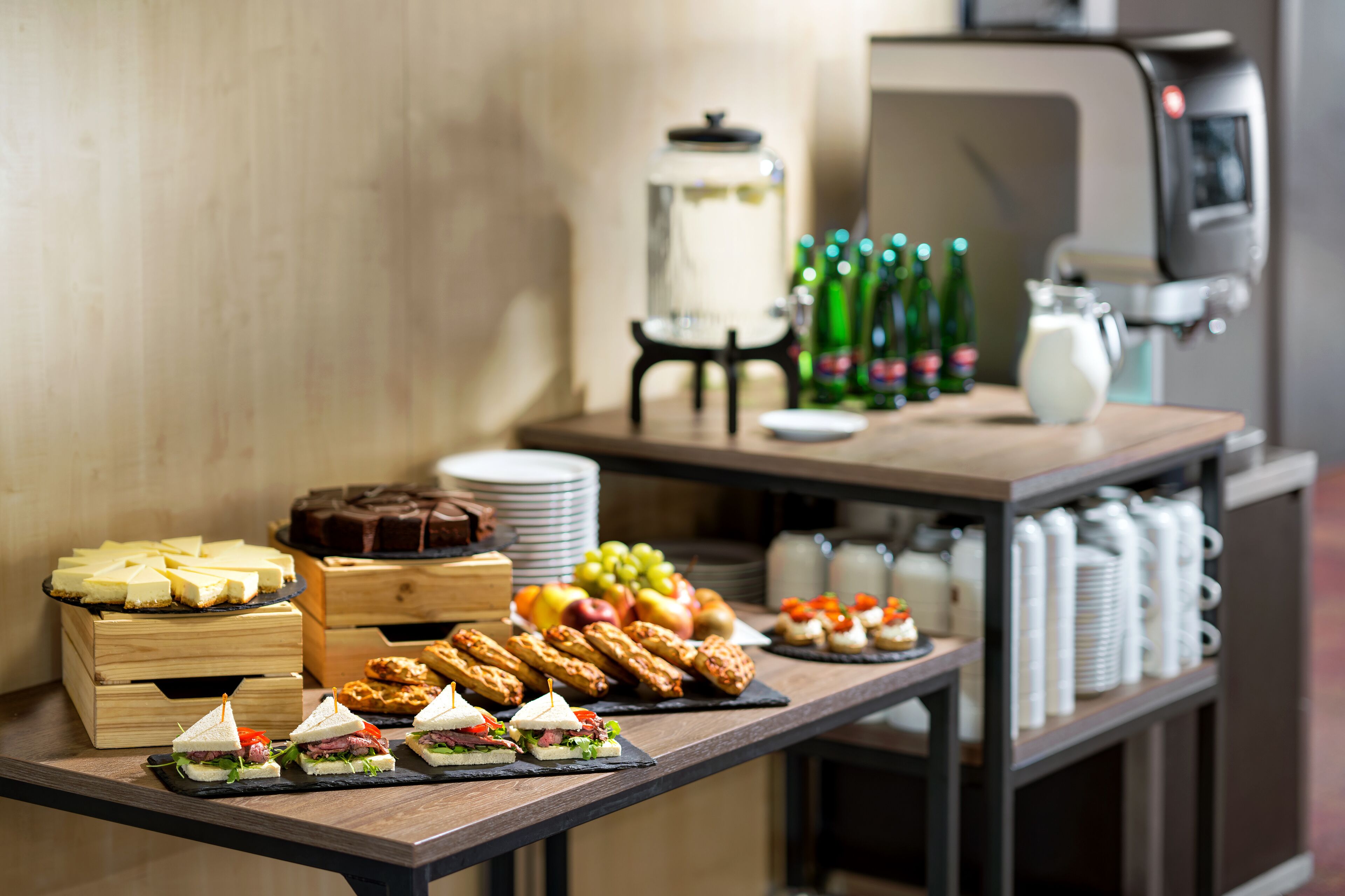 A buffet table with various food items, including sandwiches, pastries, and fruits. There is a coffee machine and bottles of soda in the background.