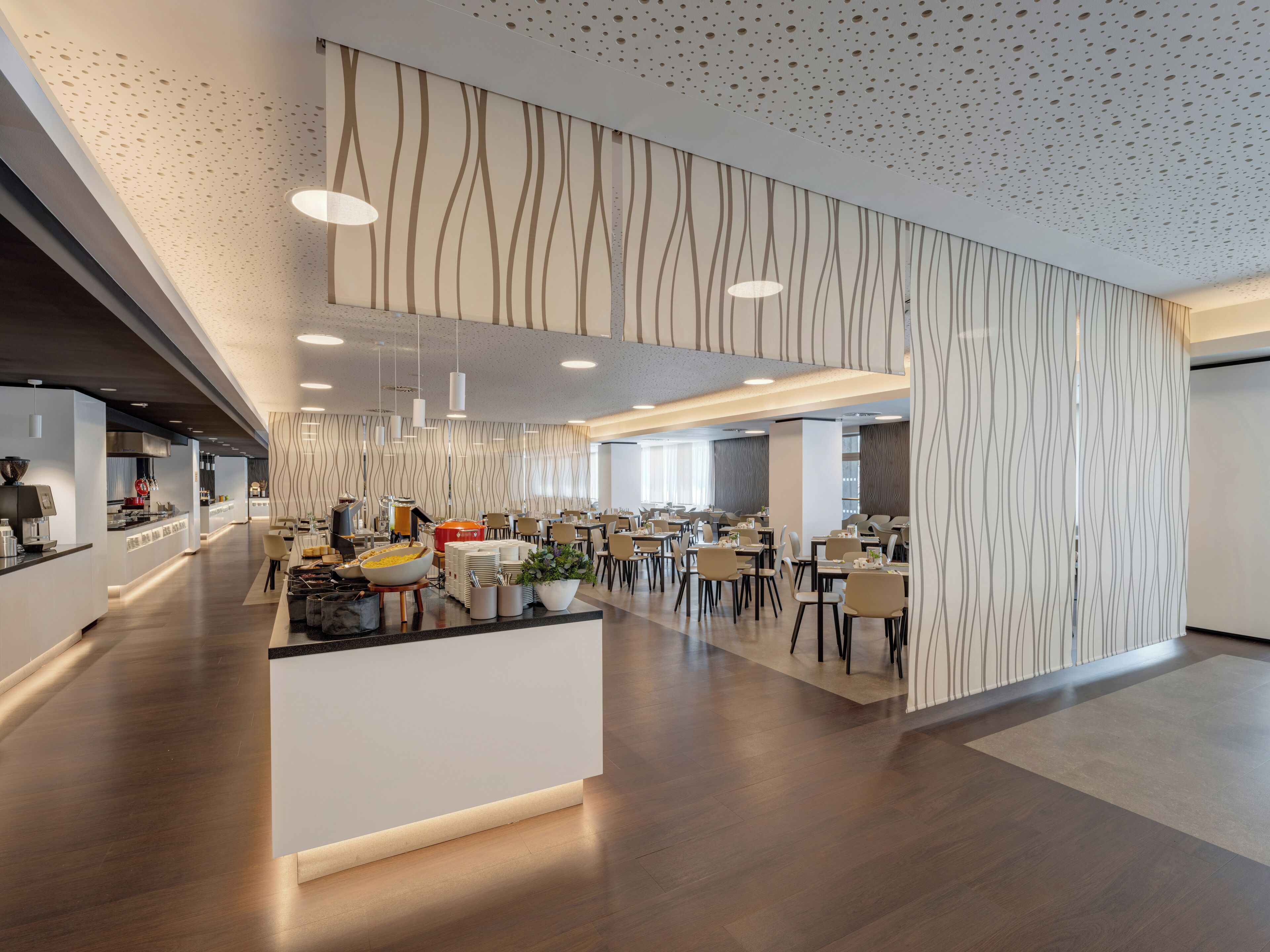 A modern hotel lobby with a central reception desk, dining area with tables and chairs, and a kitchen in the background.