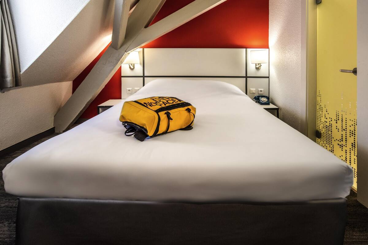 Une chambre d’hôtel avec un lit, un sac jaune, un téléphone et un mur orné d’un motif rouge et blanc.
