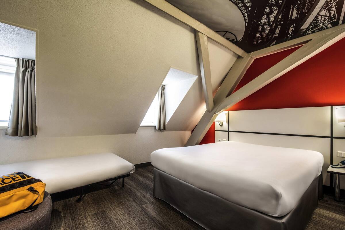 Una habitación de hotel compacta con dos camas, una mesita, una lámpara y un ventanal con cortinas.