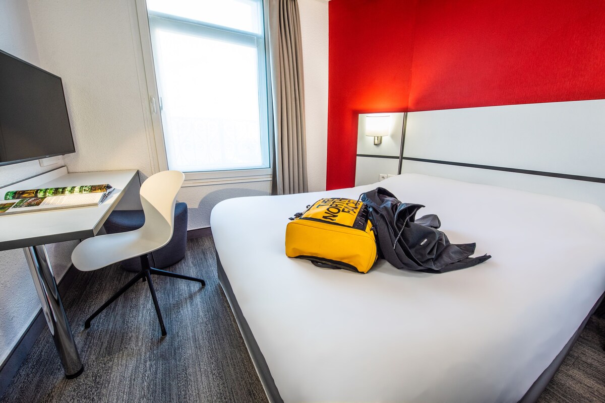 Une chambre d’hôtel avec un lit, un bureau et une chaise, un sac à dos North Face et une lampe.