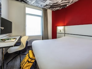 Une chambre d’hôtel avec un lit, un bureau, une chaise, une télévision, une lampe et une fenêtre avec des rideaux.