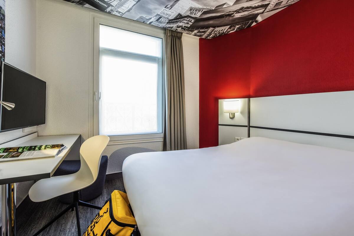 Une chambre d’hôtel avec un lit, un bureau, une chaise, une télévision, une lampe et une fenêtre avec des rideaux.