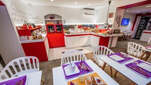 Un espace repas moderne aux couleurs blanche et rouge, avec un îlot central où sont disposés des plats et des boissons, entouré de chaises blanches et agrémenté de décorations de table violettes.
