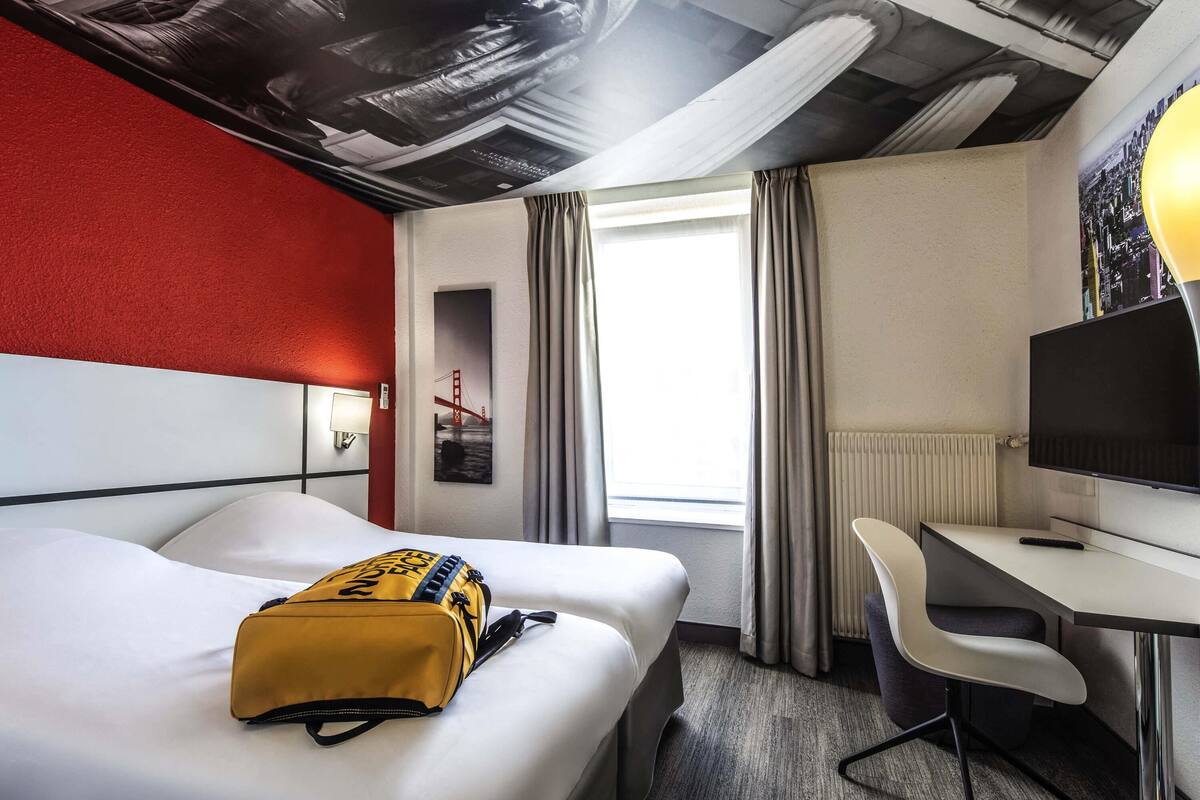 Une chambre d’hôtel comprenant un lit, un bureau, une chaise, une fenêtre avec des rideaux, un téléviseur fixé au mur et un tableau représentant un pont.