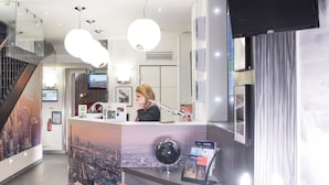 Un espace d’accueil d’hôtel au design moderne, comprenant un comptoir d’accueil, un fauteuil rouge et un luminaire décoratif.