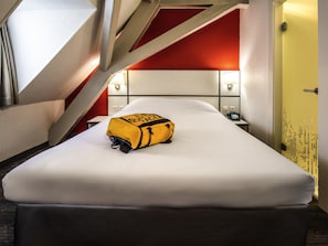 Une chambre d’hôtel avec un lit, un sac jaune, un téléphone et un mur orné d’un motif rouge et blanc.