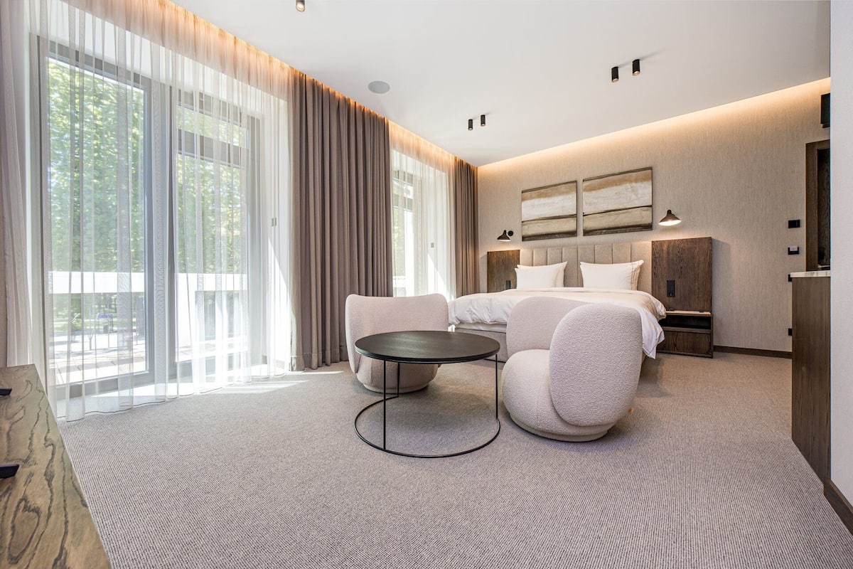 Ein modernes Hotelzimmer mit einem großen Bett, einem runden Tisch und Blick ins Freie durch große Fenster.