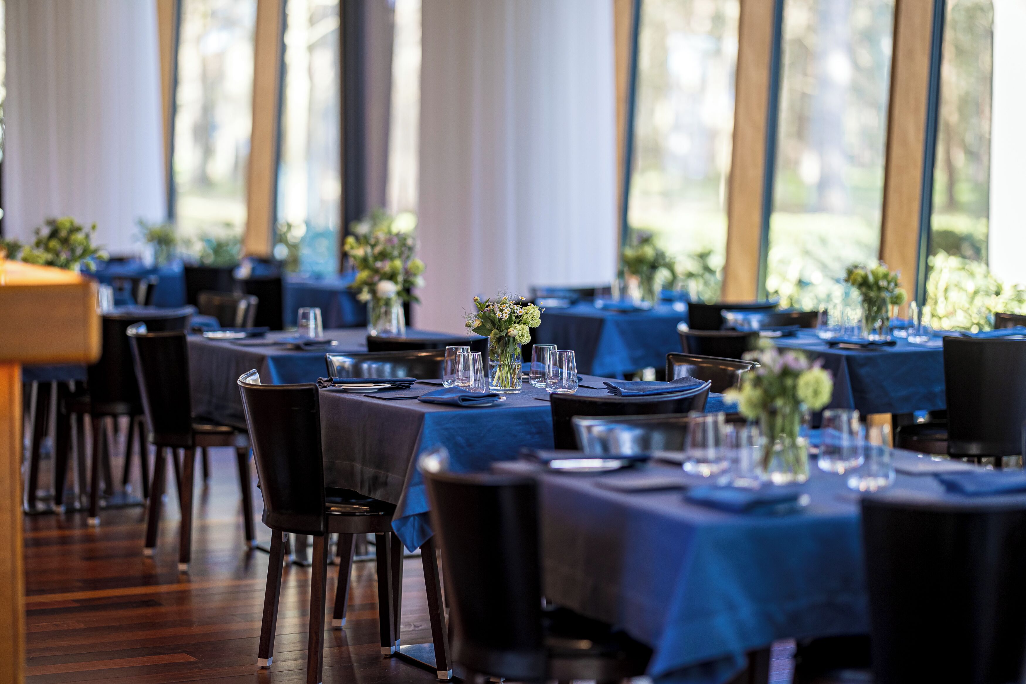 Un restaurante con varias mesas dispuestas, cubiertas con manteles azules, cristalería transparente y centros de mesa florales.