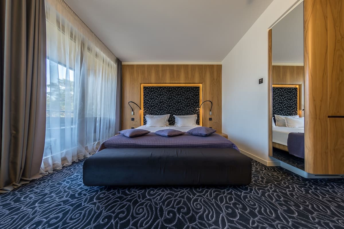 Ein modernes Hotelzimmer mit einem großen Bett, einem Holzkopfteil, einem Spiegel und Blick ins Freie.