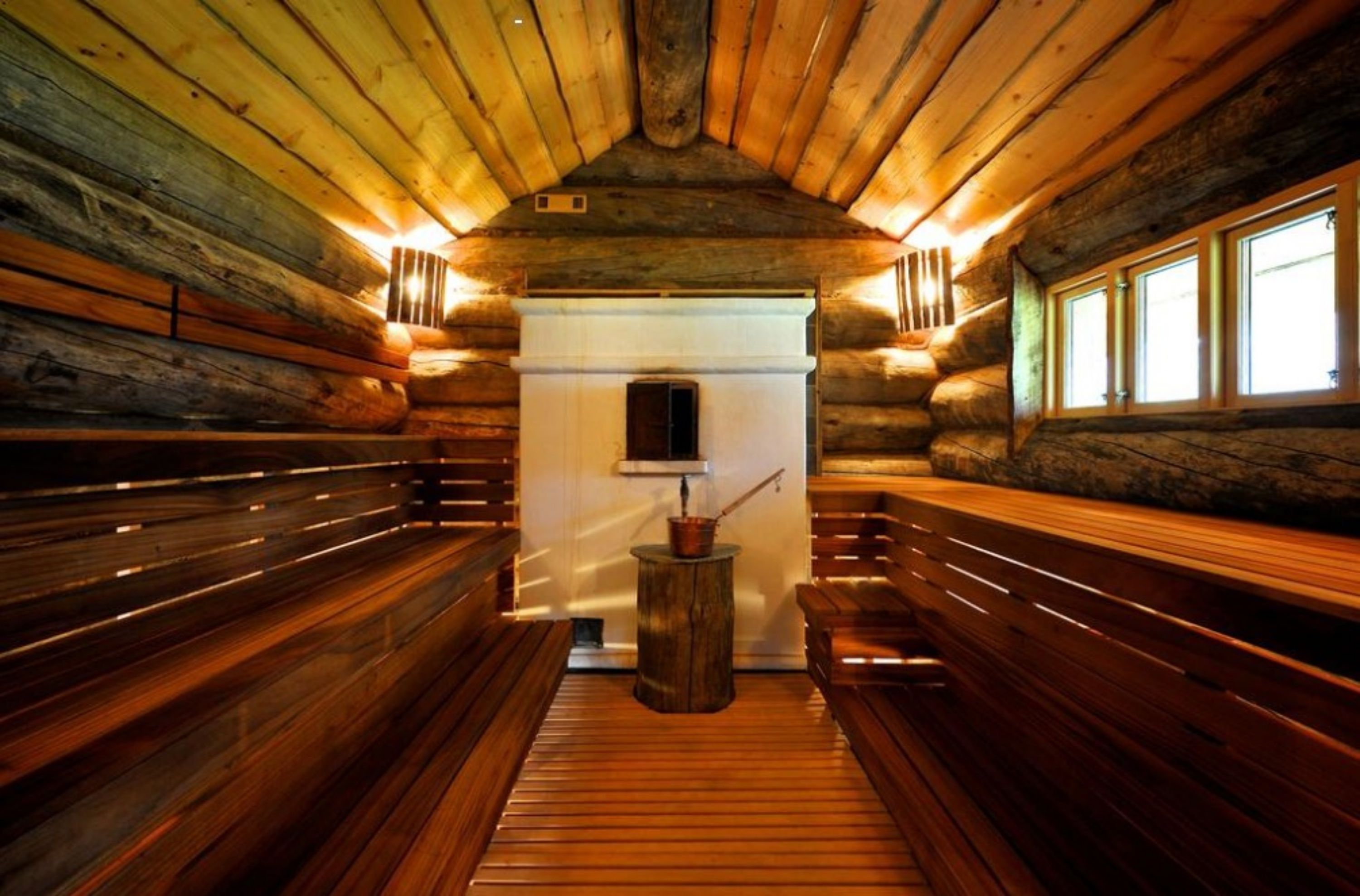Interior de una cabaña de troncos con bancos de madera y una chimenea central.