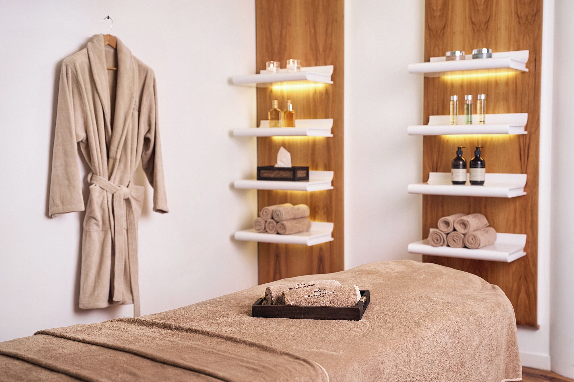 A spa room with a wooden wall, shelves with toiletries, a robe, and a massage table.