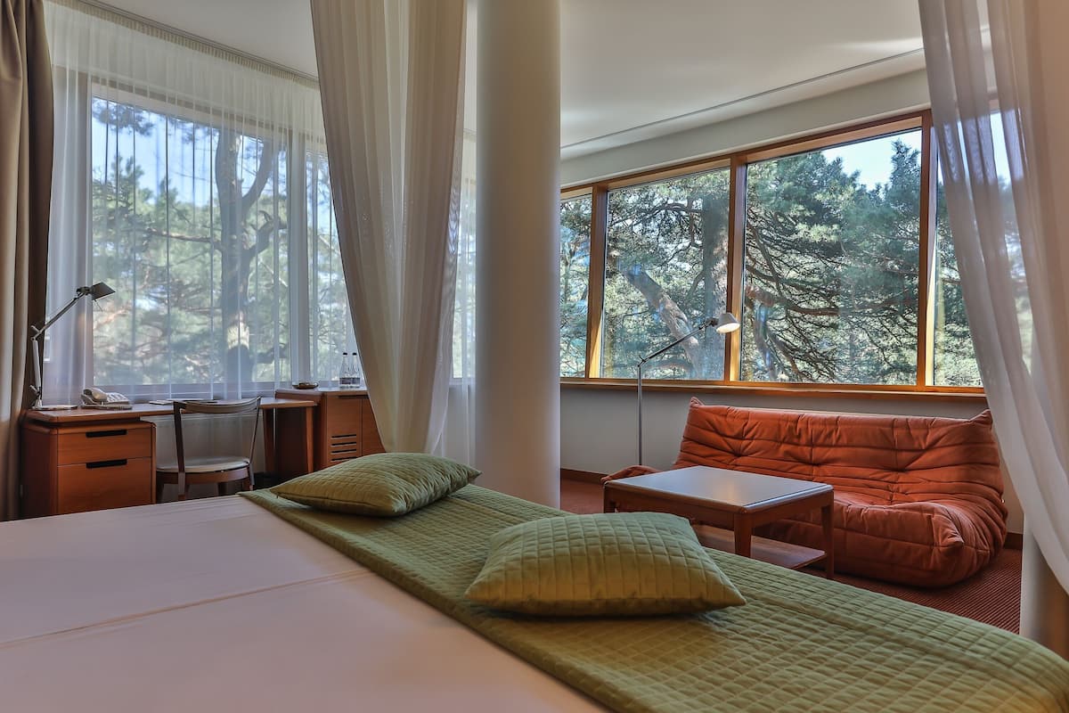Ein Schlafzimmer mit Bett, Schreibtisch, Stuhl, Sofa und Fenster mit Blick auf einen Wald.