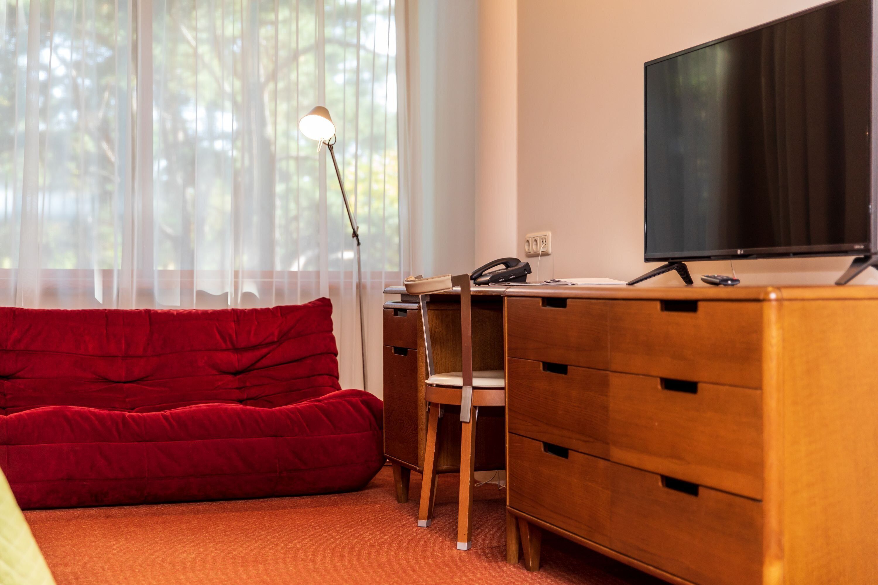 A room with a wooden desk, a chair, a red sofa, a TV, and a lamp.