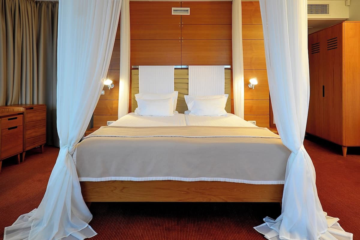 Ein Hotelzimmer mit einem großen Bett, weißen Vorhängen, einem Holz-Kopfbrett, Nachttischlampen und einem Schrank aus Holz.