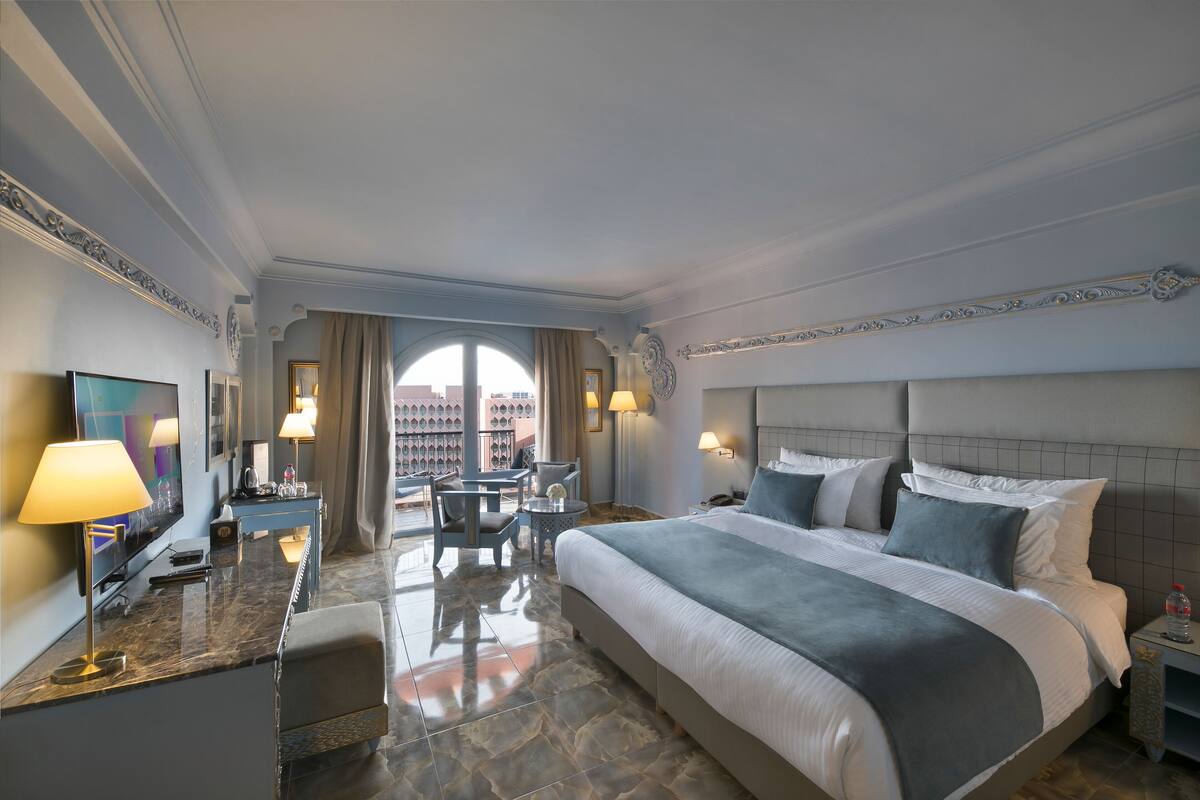 A hotel room with a large bed, a desk with a lamp, a television, and a view of the city through a large window.