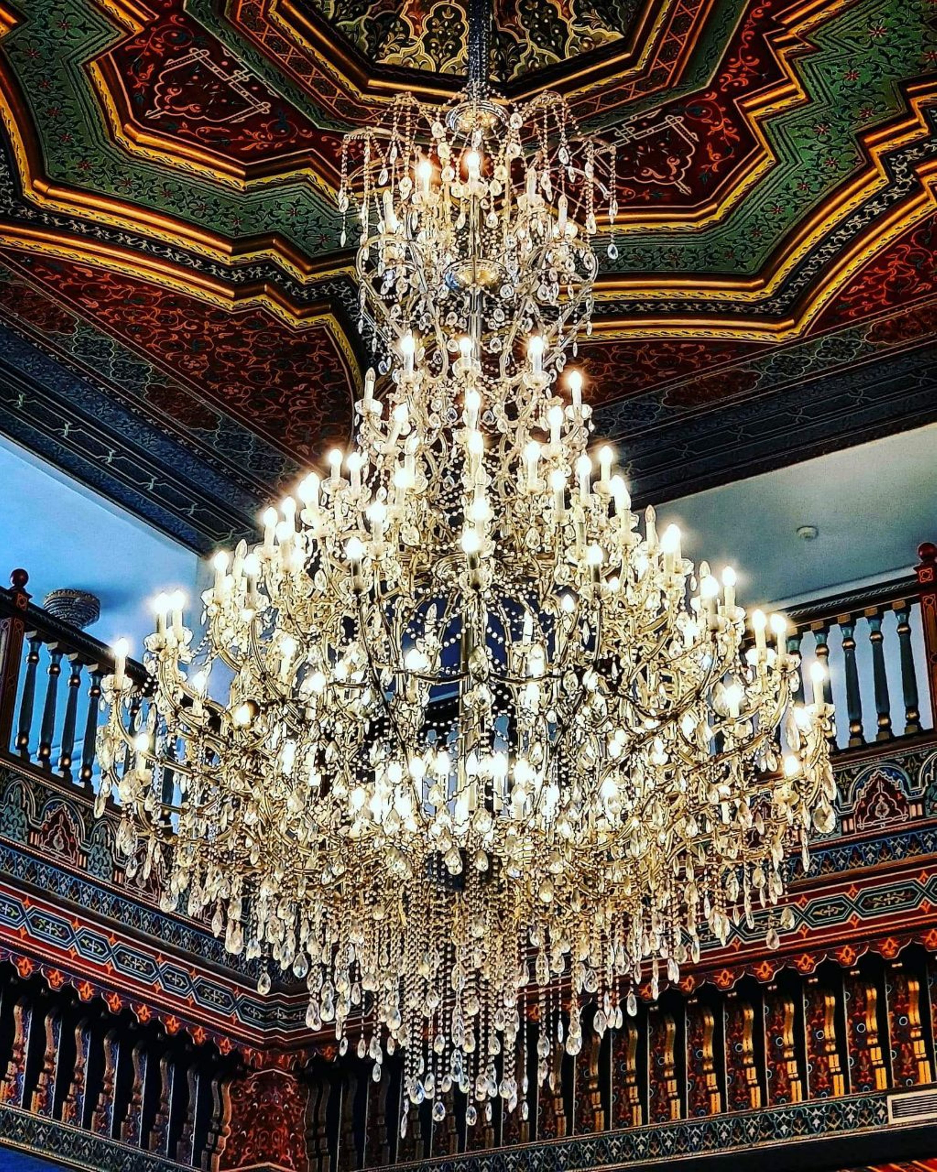 A grand chandelier with multiple tiers and candle-like lights, set against a richly decorated ceiling with geometric patterns and a balcony railing visible in the background.
