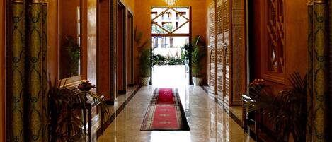 A grand hallway with ornate columns, chandeliers, and a red carpet leading to a bright interior.