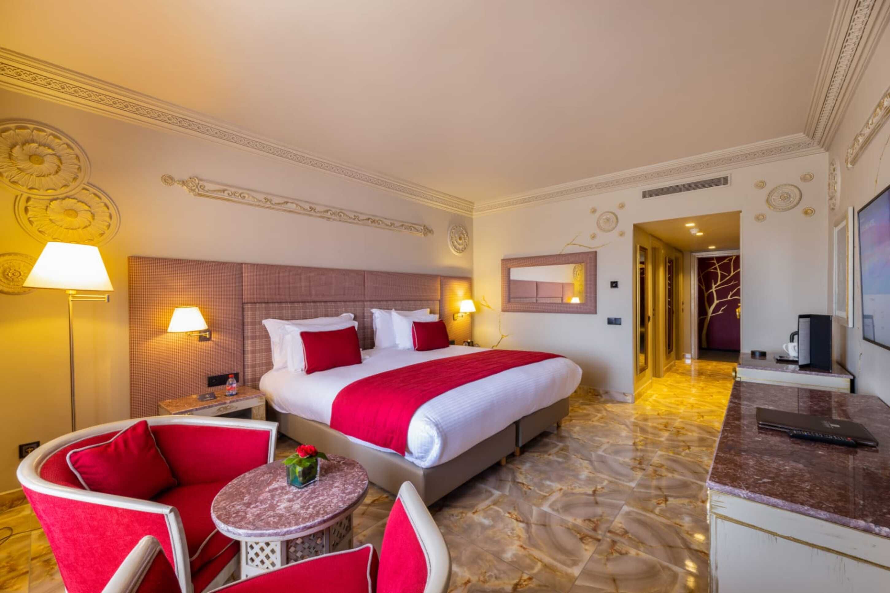 A hotel room with a large bed, red and white bedding, a marble floor, a small round table with a vase, and a TV on a marble counter.