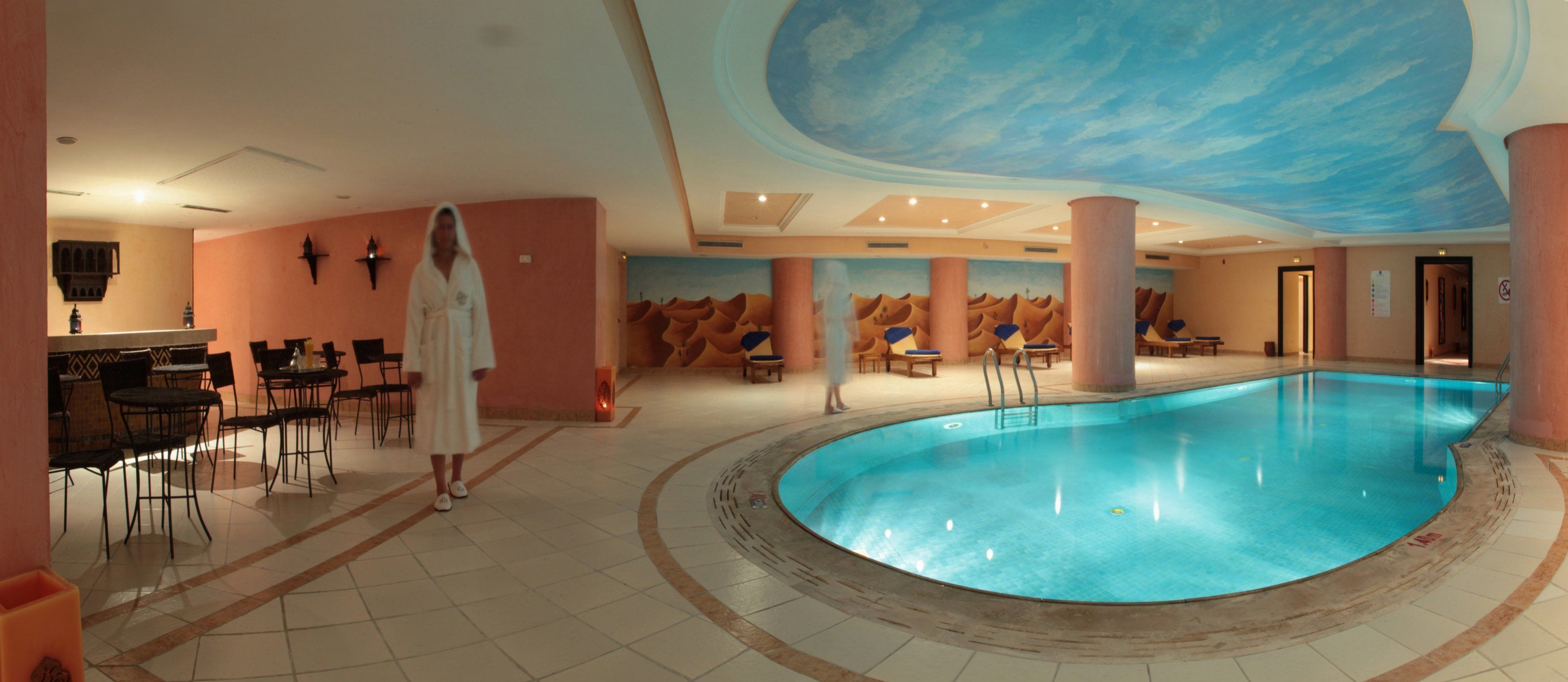 Una piscina interior con un mural azul en el techo, una zona de estar con sillas y mesas, y una persona vestida con bata.