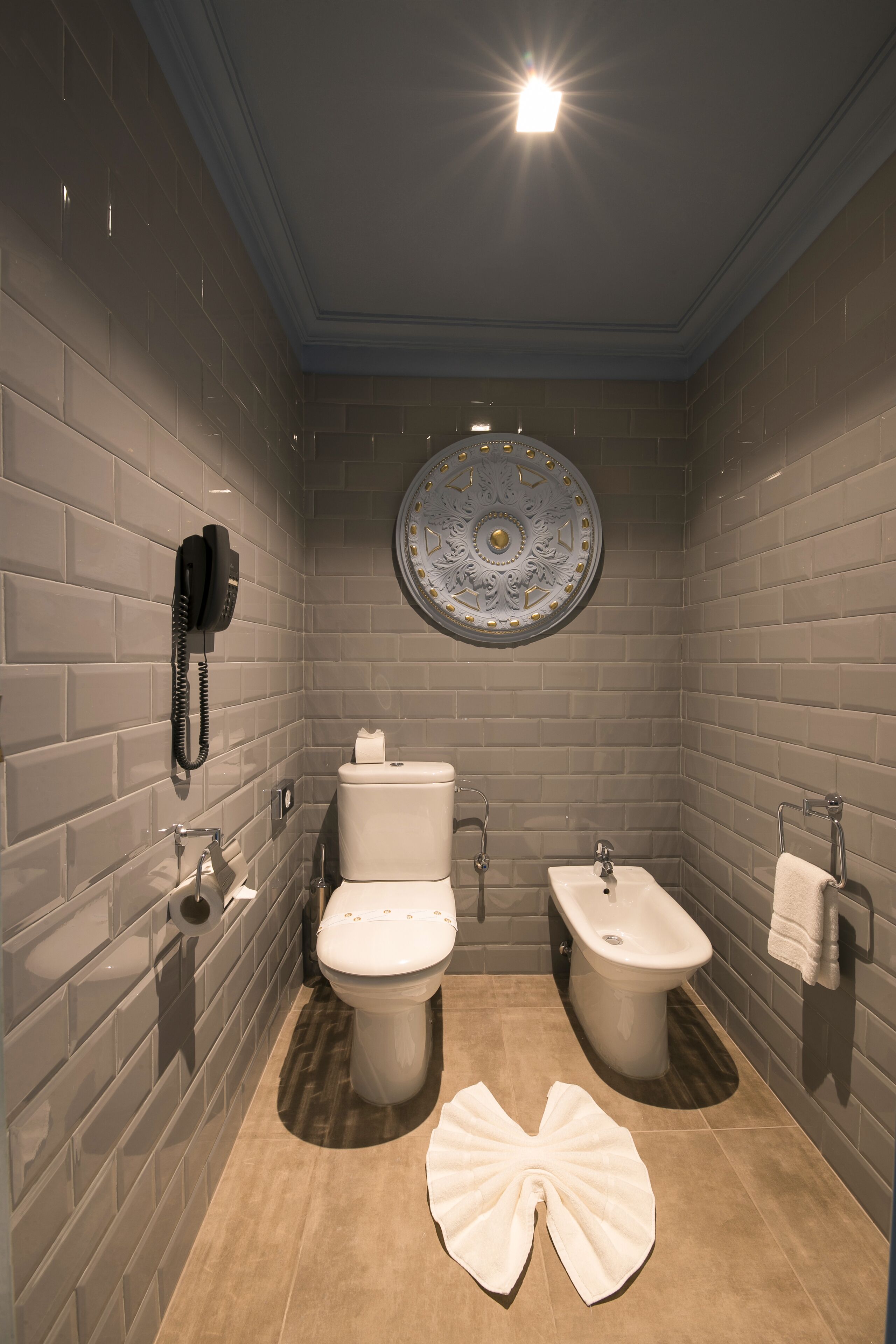 A modern bathroom with grey tiled walls, a round decorative mirror, a toilet, a bidet, and a towel on the floor.