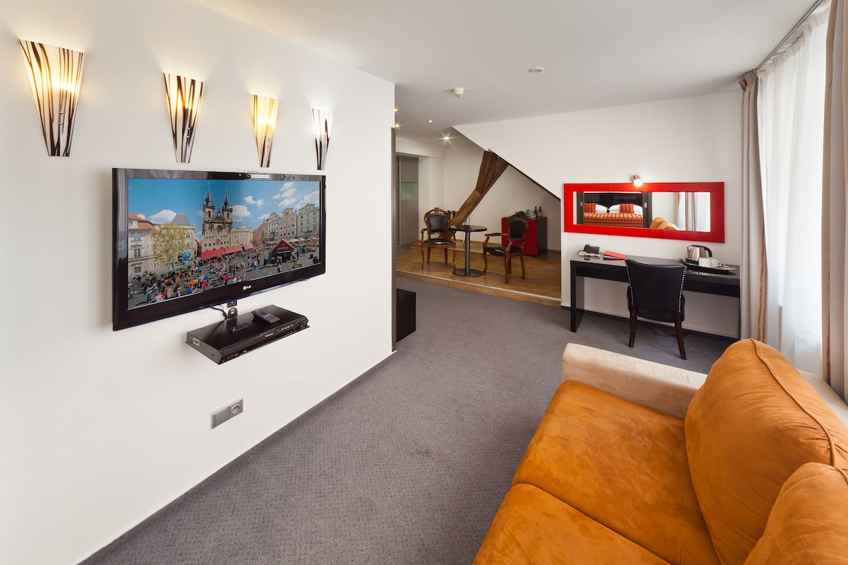 Una habitación de hotel moderna con un televisor de pantalla plana grande montado en la pared, un sofá naranja cómodo, un escritorio con una silla, y una escalera que lleva a un nivel superior.