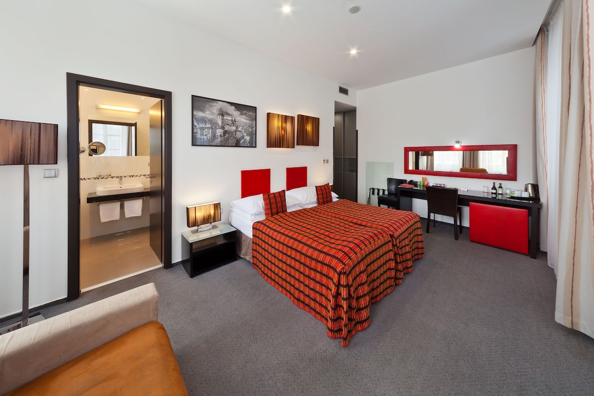 Habitación de hotel con cama, escritorio, silla, espejo y baño visible a través de una puerta abierta.