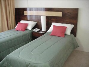A hotel room with two single beds, each with a green bedspread and a red pillow. There is a wooden headboard with a light fixture above the beds.