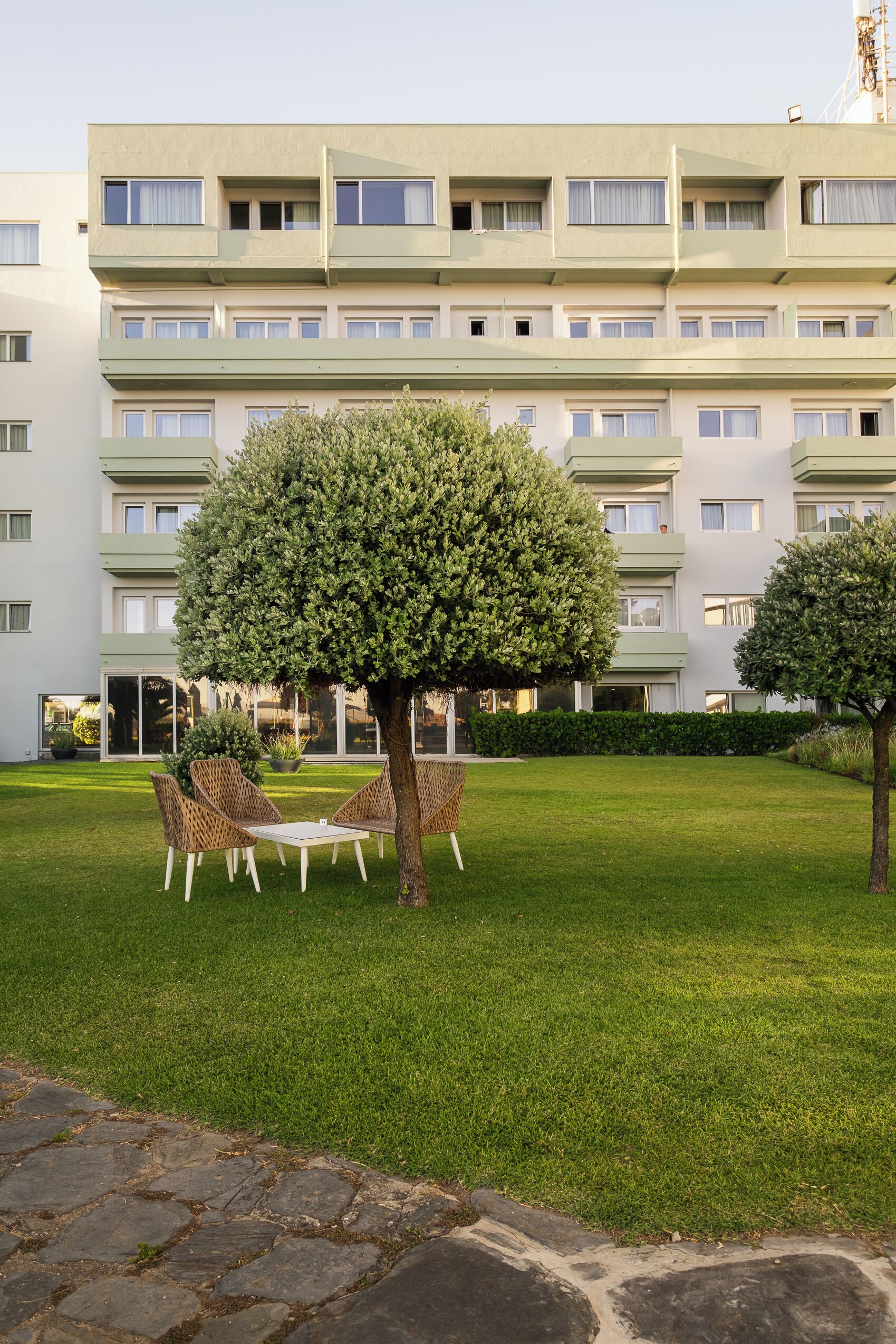 Un hotel de varios pisos con fachada verde, césped cuidado y zona de estar al aire libre.