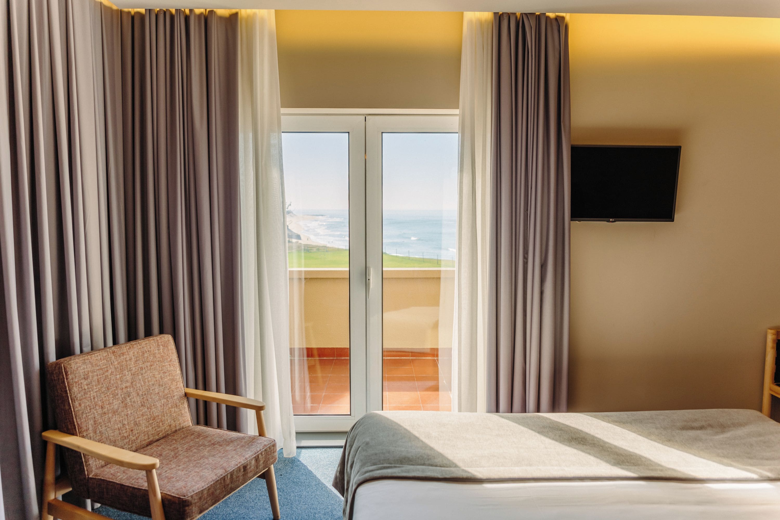 Une chambre d’hôtel avec un lit, une chaise, une télévision et une vue sur l’océan par une porte vitrée.
