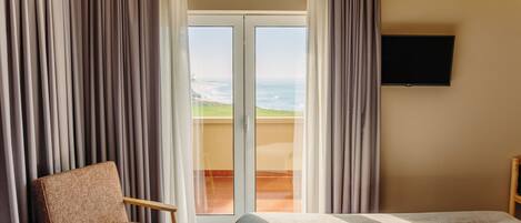Une chambre d’hôtel avec un lit, une chaise, une télévision et une vue sur l’océan par une porte vitrée.
