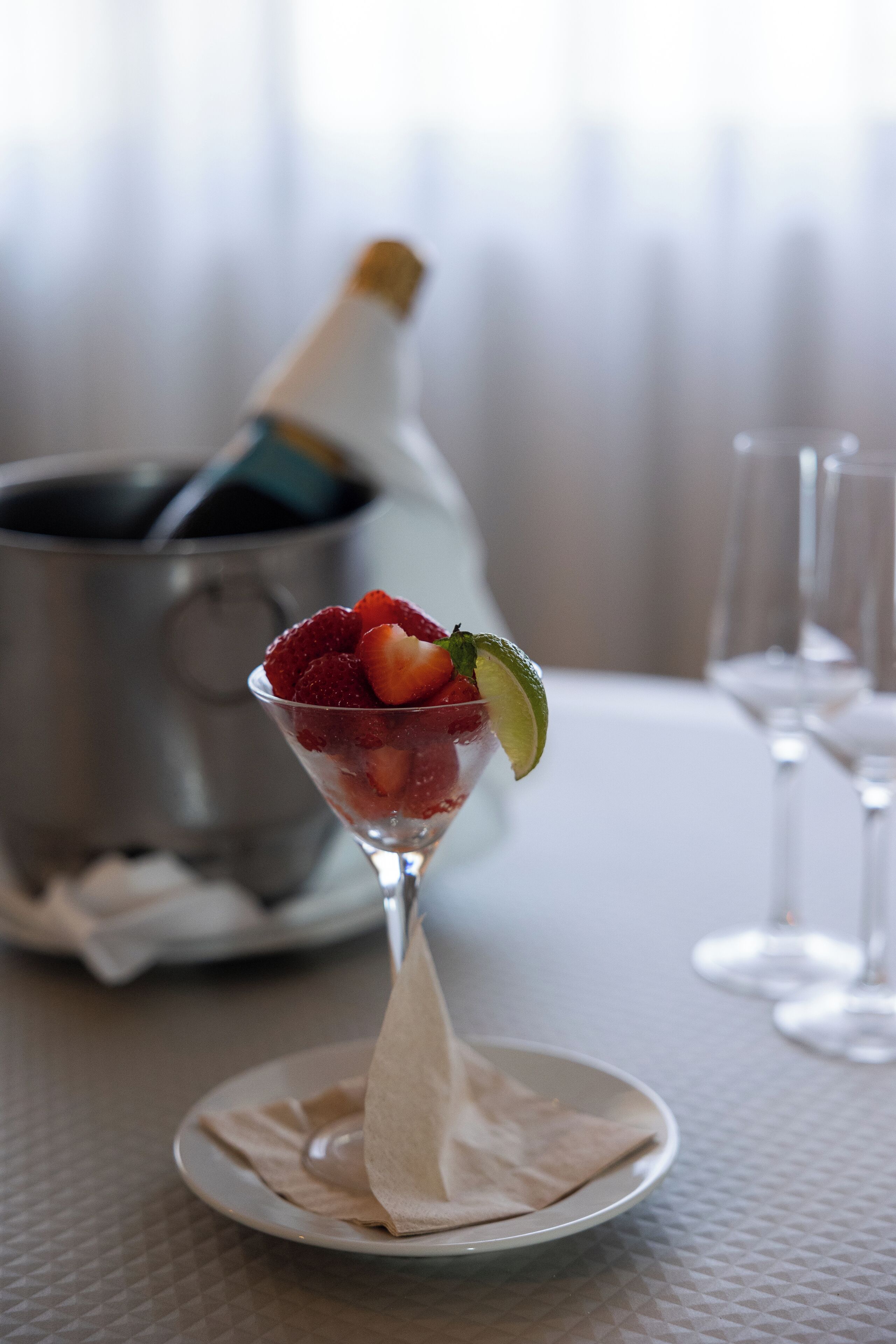 A martini glass with a strawberry and lime garnish on a white plate, a bottle of champagne, and a champagne bucket in the background.