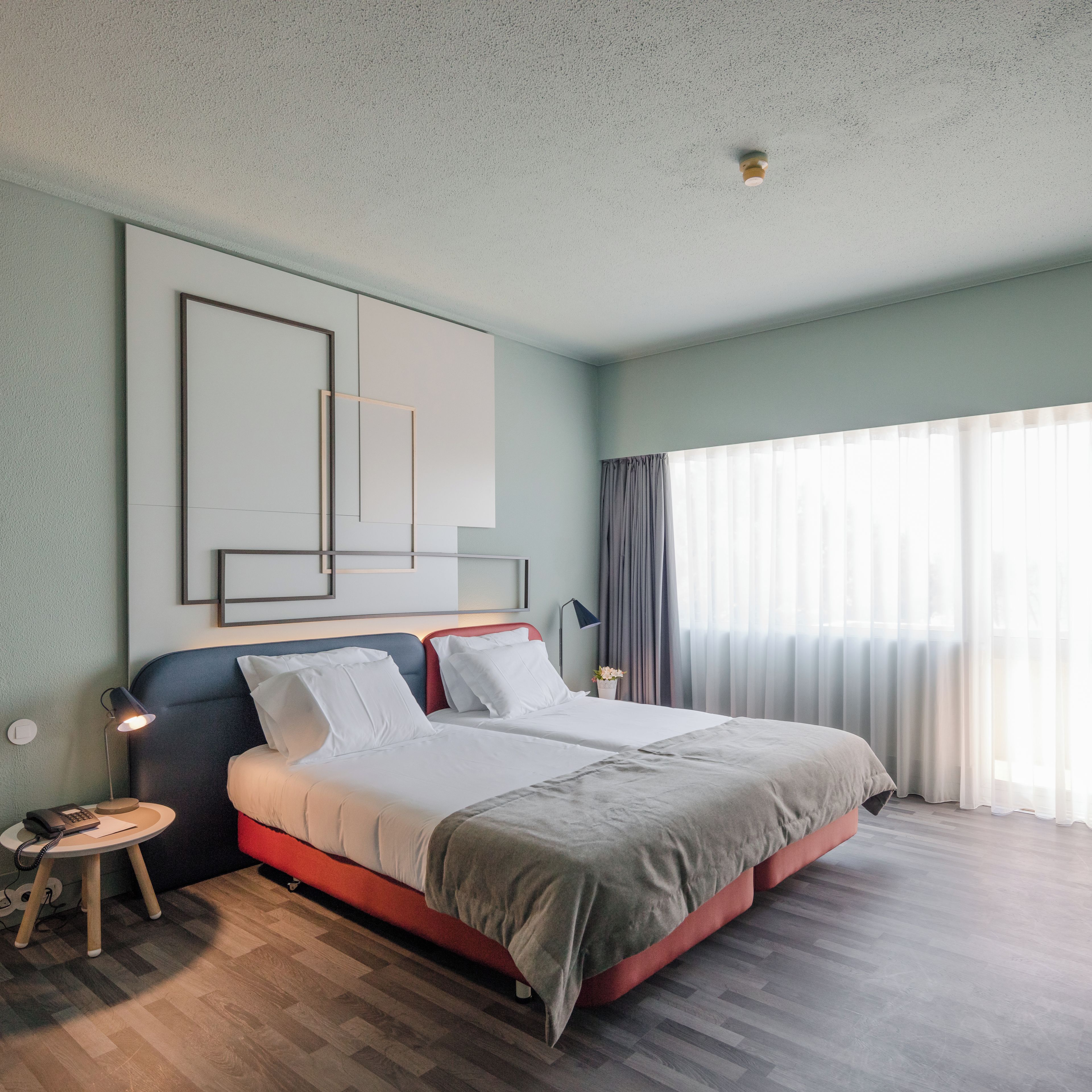 Une chambre d’hôtel moderne avec un grand lit, une table de chevet avec une lampe et une vue sur l’extérieur à travers des rideaux légers.