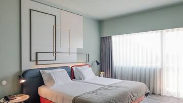 Une chambre d’hôtel moderne avec un grand lit, une table de chevet avec une lampe et une vue sur l’extérieur à travers des rideaux légers.