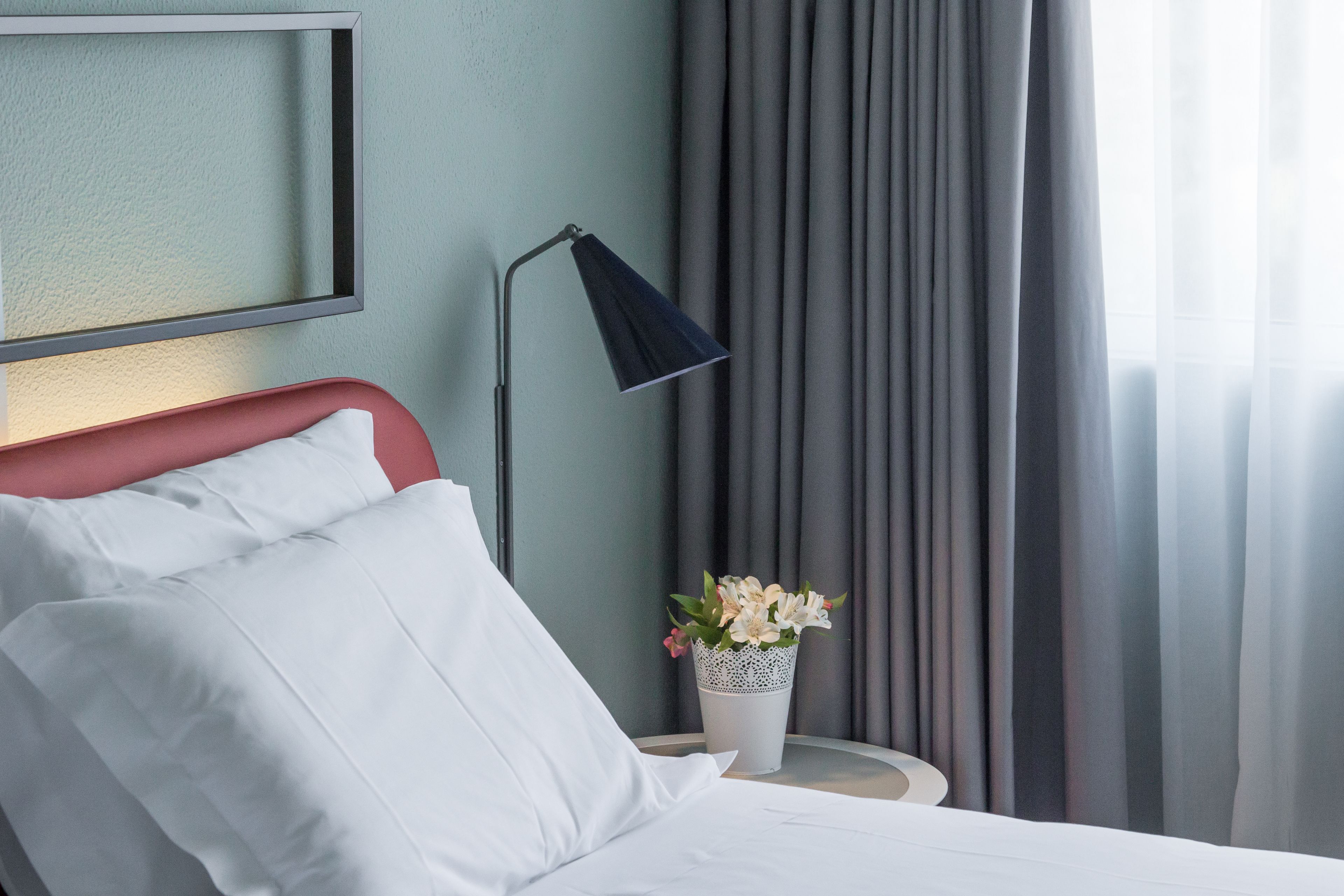 A neatly made bed with white linens, a red headboard, a bedside lamp, and a small vase with flowers on a bedside table.