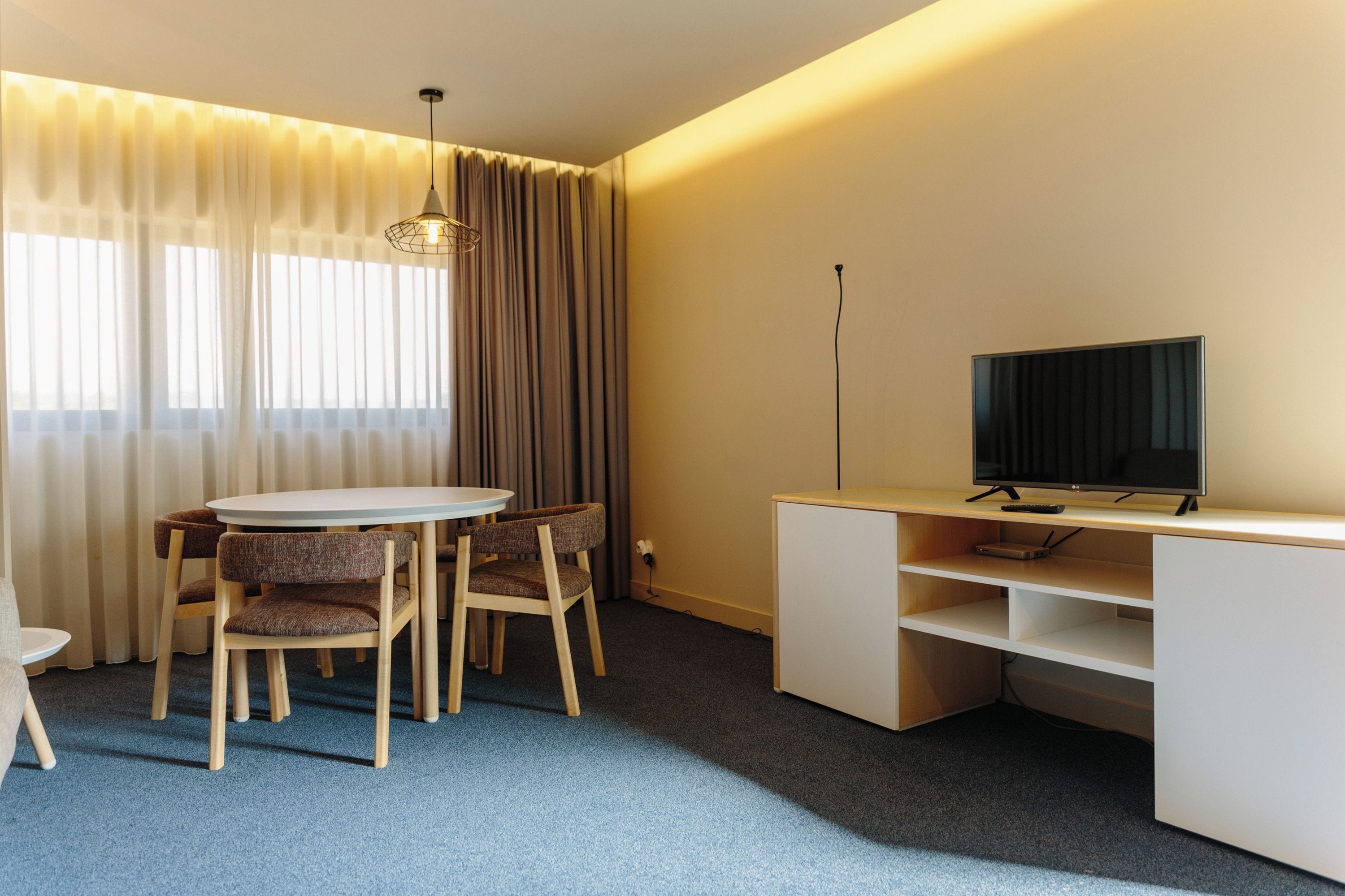 A hotel room with a round table, two chairs, a television on a stand, and a desk with shelving.
