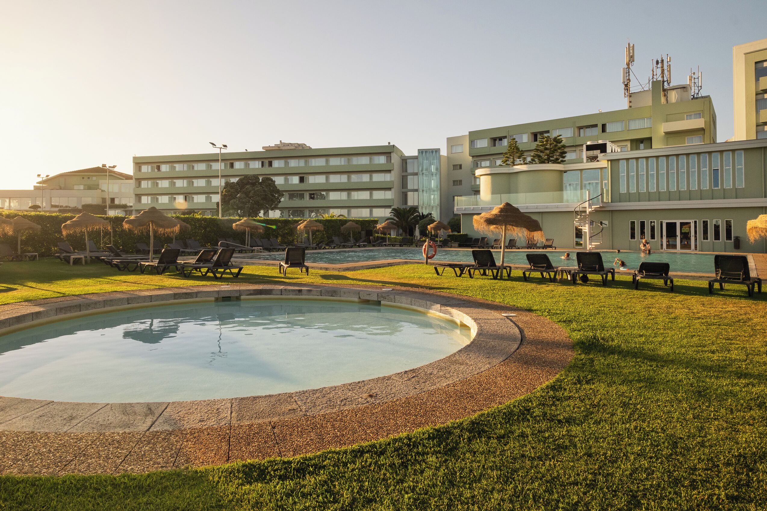 Un área de piscina con sillones de descanso y sombrillas, rodeada de césped cuidado y un edificio con varios pisos y ventanas.