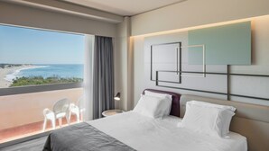 A hotel room with a large window offering a view of the beach, a bed with white linens, a modern wall art piece, and a small table with a lamp. - Axis Ofir Beach Resort Hotel (Esposende)