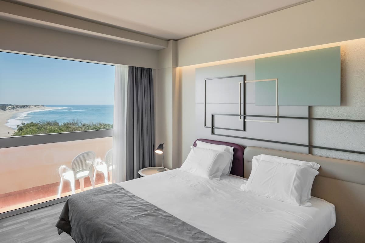 Habitación de hotel con un ventanal grande que ofrece vistas a la playa, una cama con ropa de cama blanca, una obra de arte moderna en la pared y una pequeña mesa con lámpara.