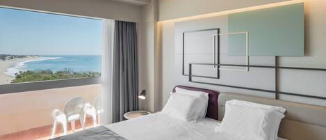 Habitación de hotel con un ventanal grande que ofrece vistas a la playa, una cama con ropa de cama blanca, una obra de arte moderna en la pared y una pequeña mesa con lámpara.