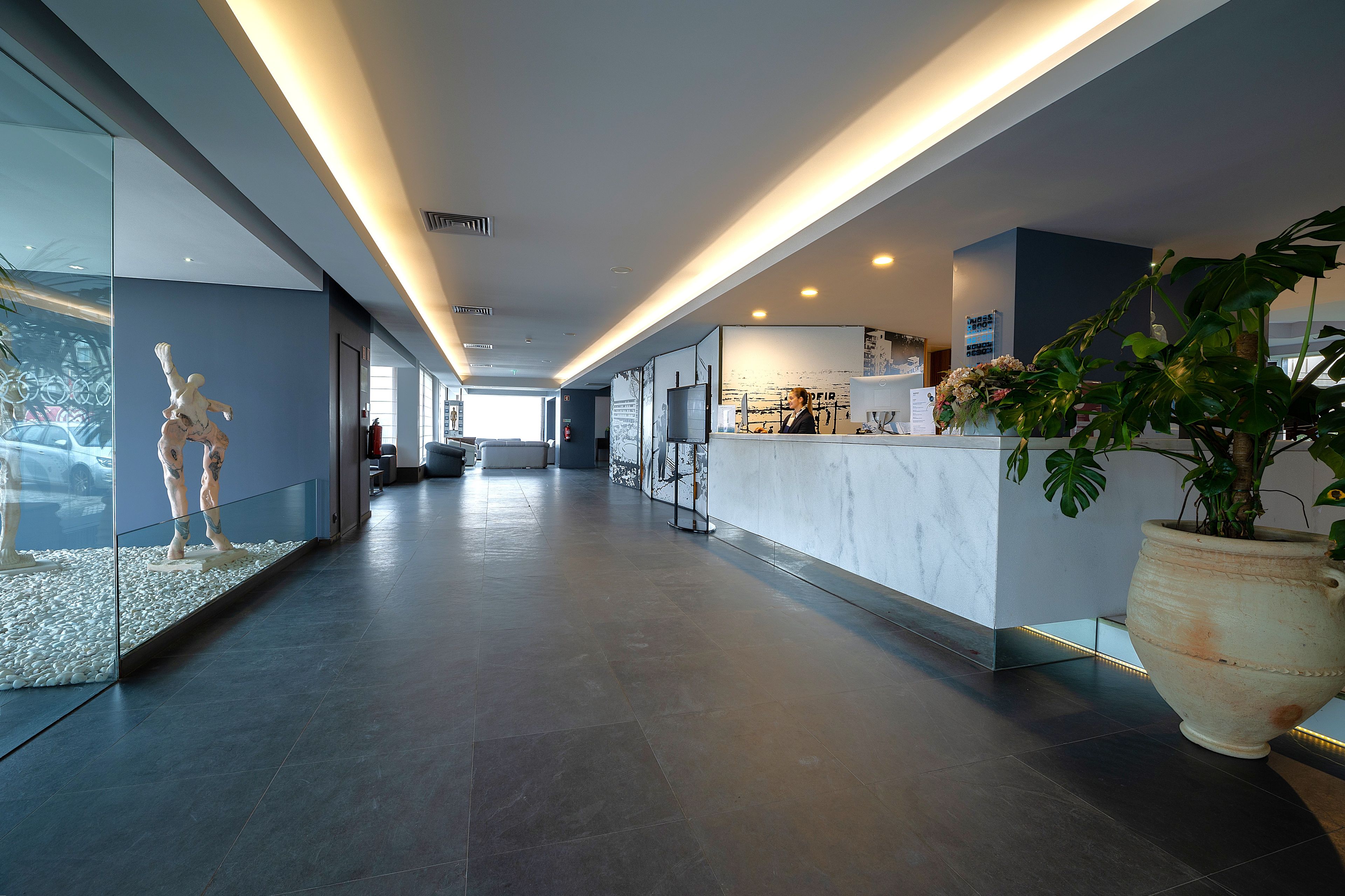 A modern hotel lobby with a reception desk, a large potted plant, and a sculpture on display.