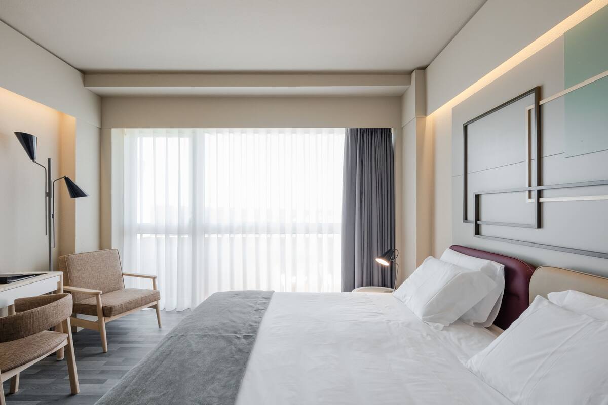 Habitación de hotel con cama, escritorio, dos sillas, lámpara y un amplio ventanal con cortinas.