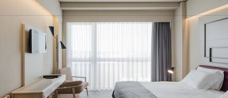 A hotel room with a bed, desk, chair, TV, and large window with curtains.