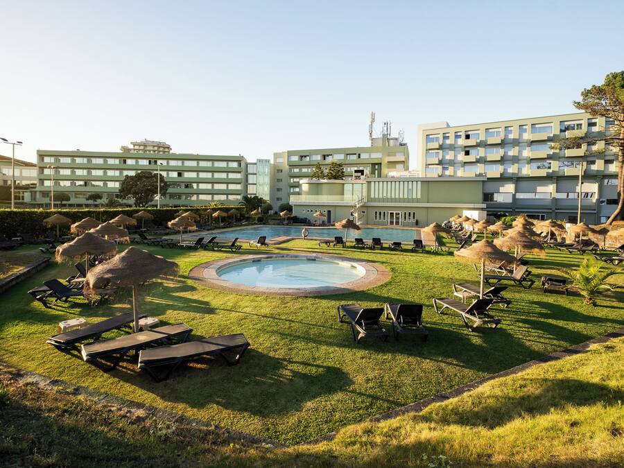 Zona de piscina con tumbonas y sombrillas, rodeada por un césped cuidado y un edificio con varios balcones.