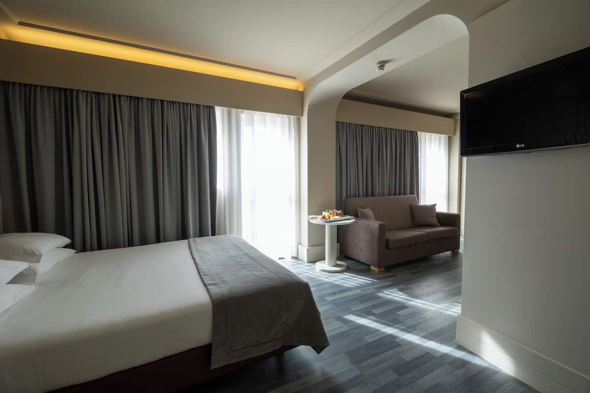 Habitación de hotel con cama, sofá, una mesita con una bandeja de frutas y un televisor montado en la pared.