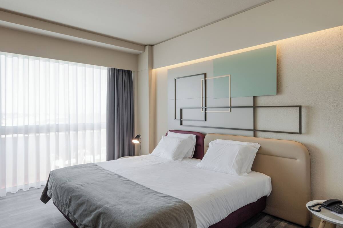 A hotel room with a bed, a bedside table with a phone, a wall-mounted light, and a window with curtains.