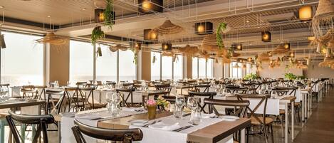 A spacious restaurant with multiple tables and chairs, large windows, and hanging plants.