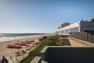 Un hotel frente a la playa con diseño moderno, que cuenta con un largo camino que lleva a la arena y un edificio con grandes ventanales con vistas al océano.